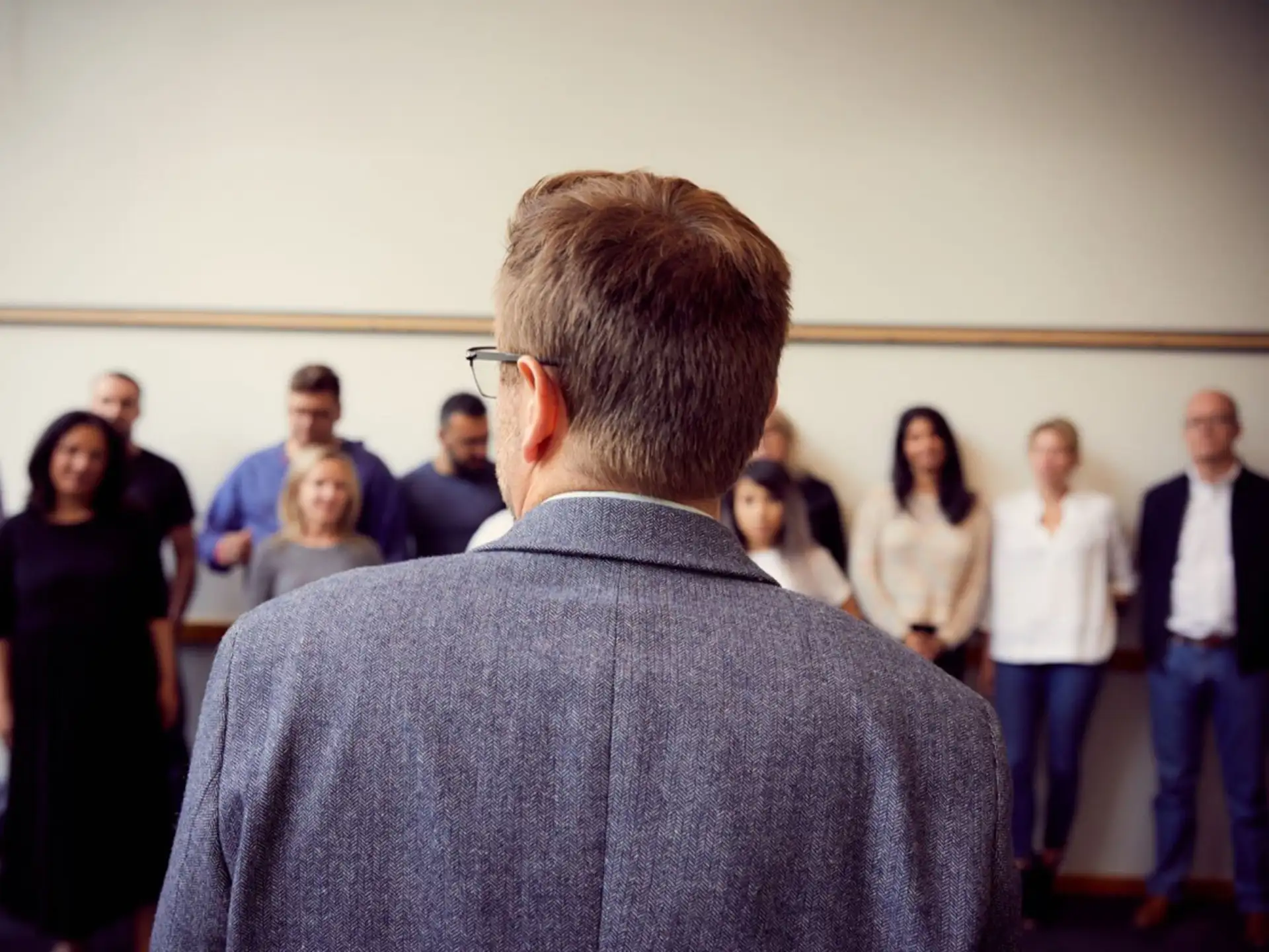 Man in a grey blazer, seen from behind, addressing a group of people standing along a wall.