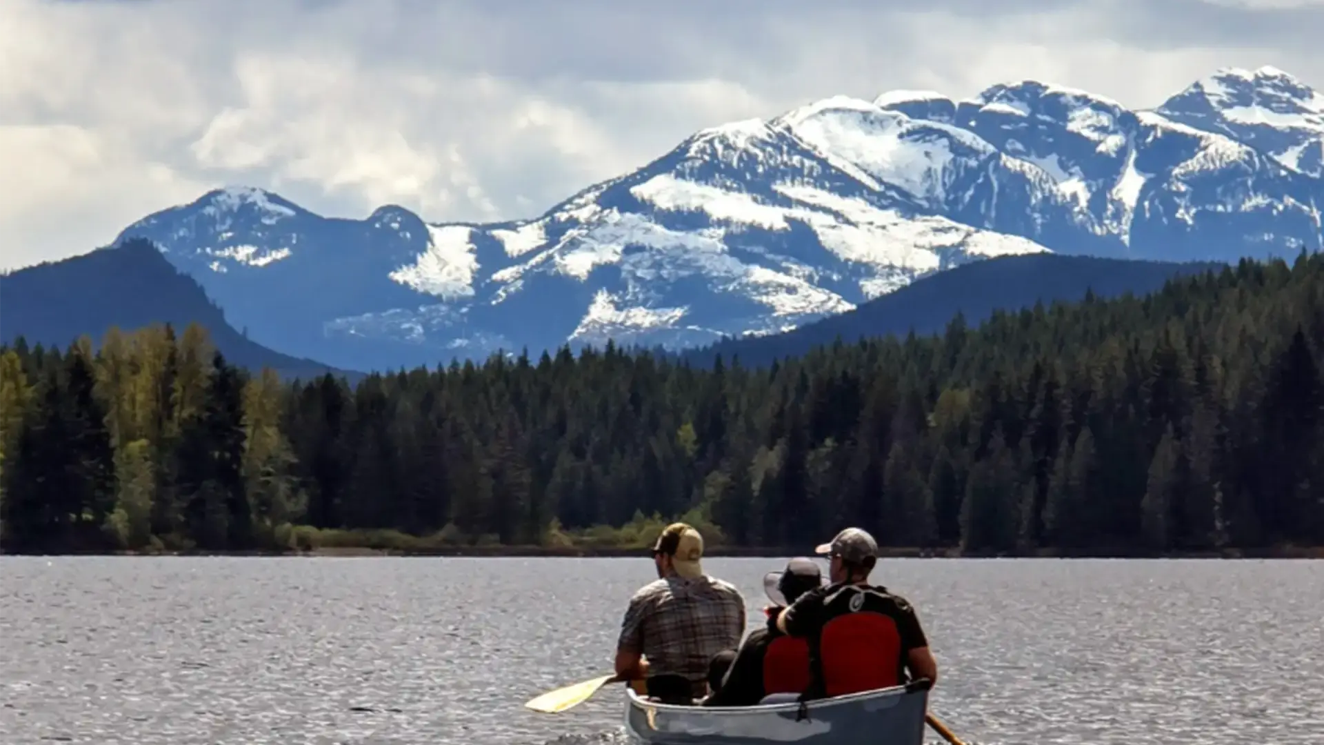Three people paddling a canoe across a lake toward a forested shoreline, with mountains in the distance.