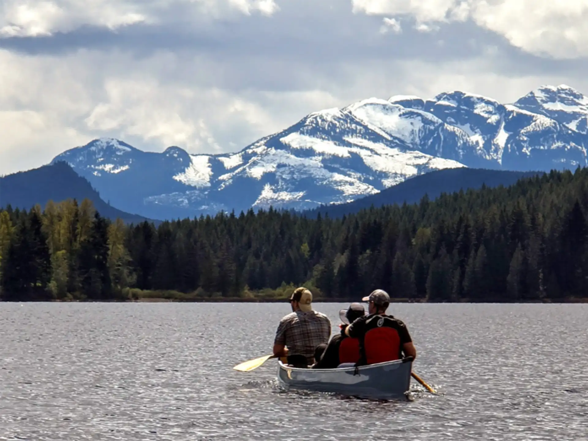 Three people paddling a canoe across a lake toward a forested shoreline, with mountains in the distance.