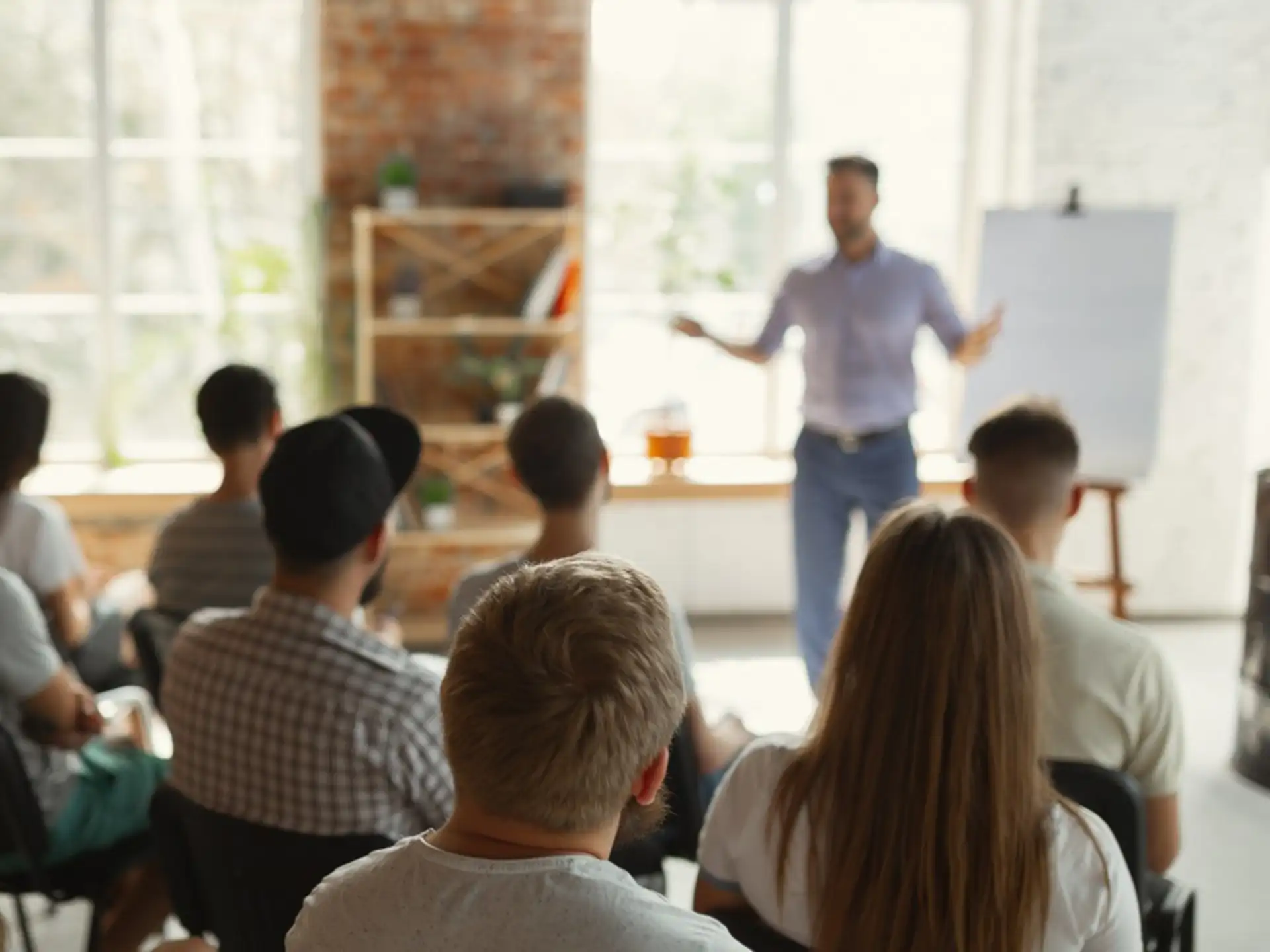 Man presenting to a seated audience in a bright, brick-walled workshop space.