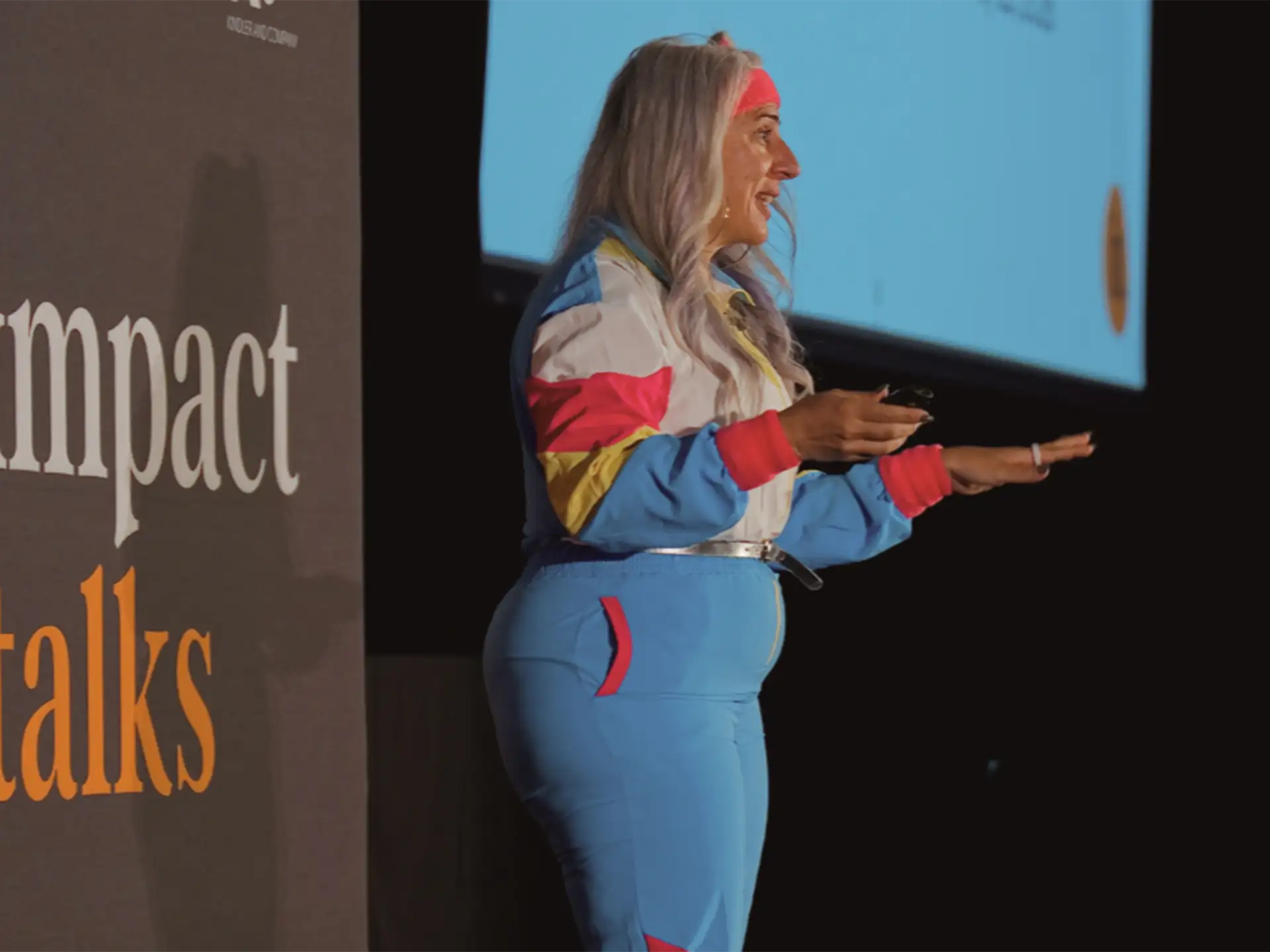 Zayna Khayat speaking onstage at Impact Talks in a colourful 1980s-style tracksuit and headband.